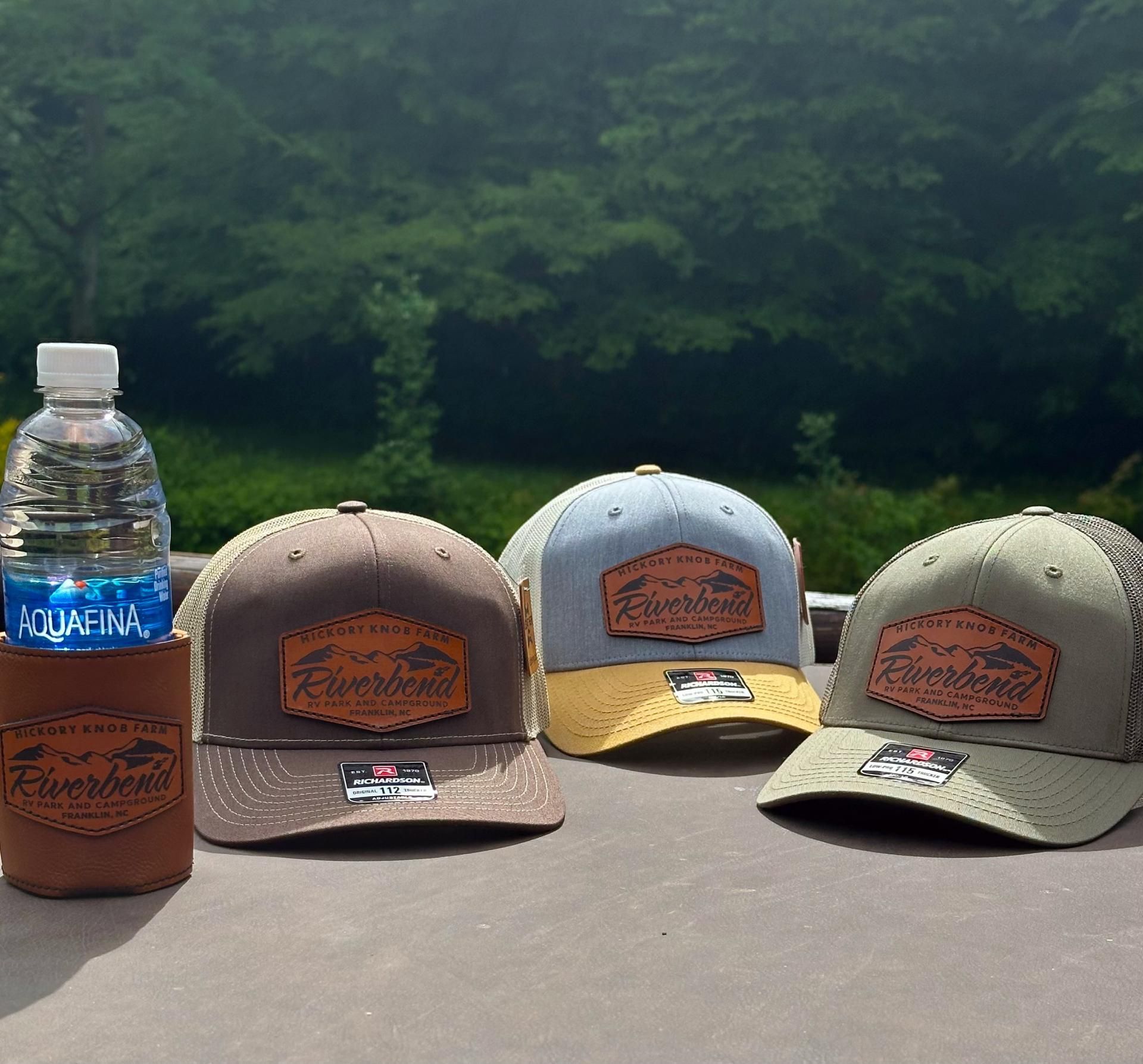 Three hats and a water bottle with a hat koozie on a tabletop outdoors. The hats are brown, yellow, and green, all with patches.
