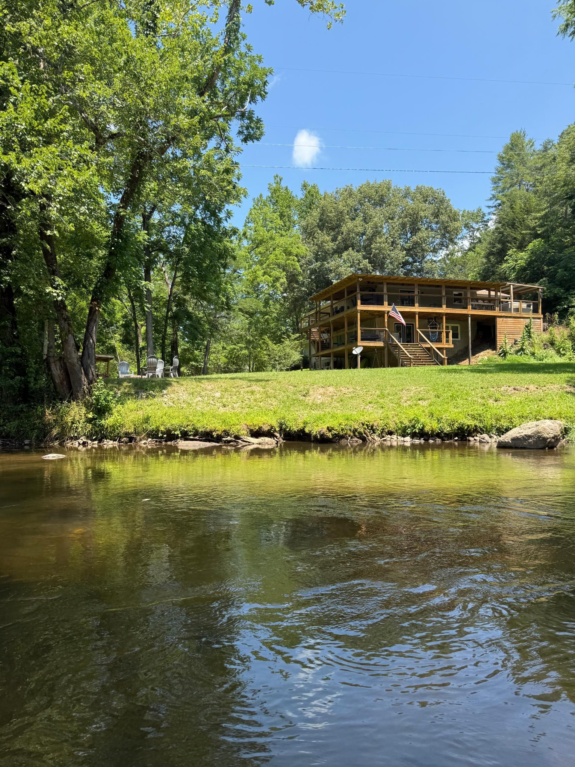River view of a multi-level wooden house with a deck nestled amongst green trees under a blue sky.