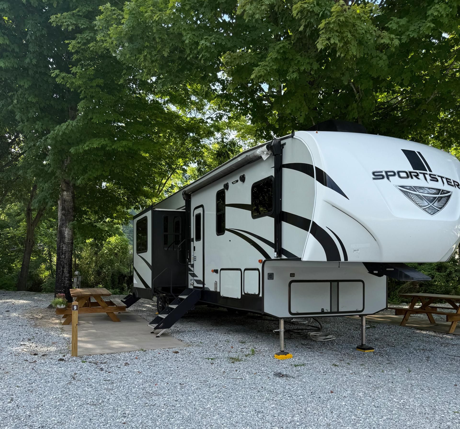 White camper trailer parked at a campsite on gravel with picnic tables and trees.