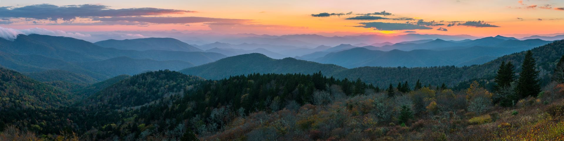 Mountain range at sunset, layered blue peaks with trees in the foreground and pink/orange sky.