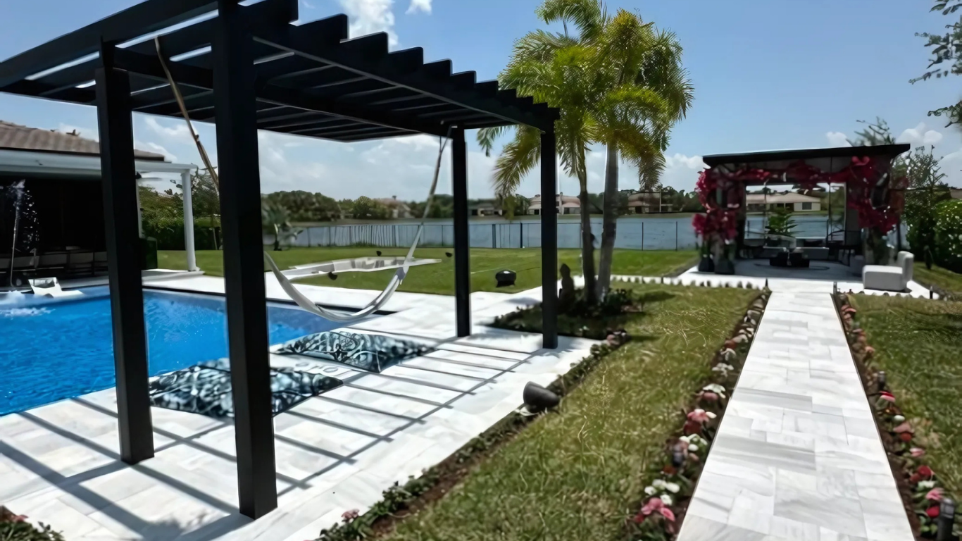 Backyard with a pool, pergola, and path lined with flowers. Palm trees and clear sky.