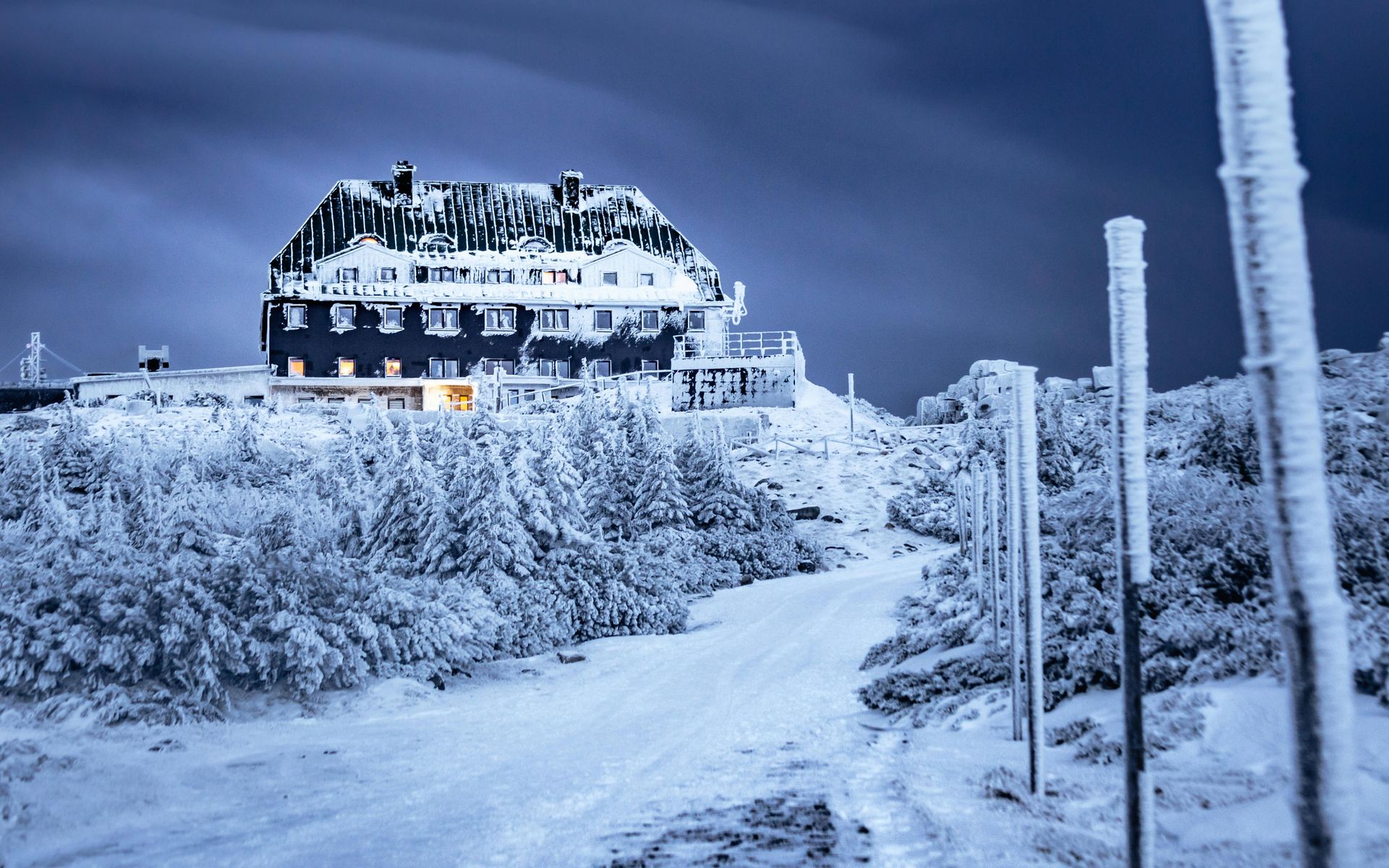 Snow-covered building on a hilltop. Winter scene with frosted trees and a path leading to the lit structure under a blue sky.