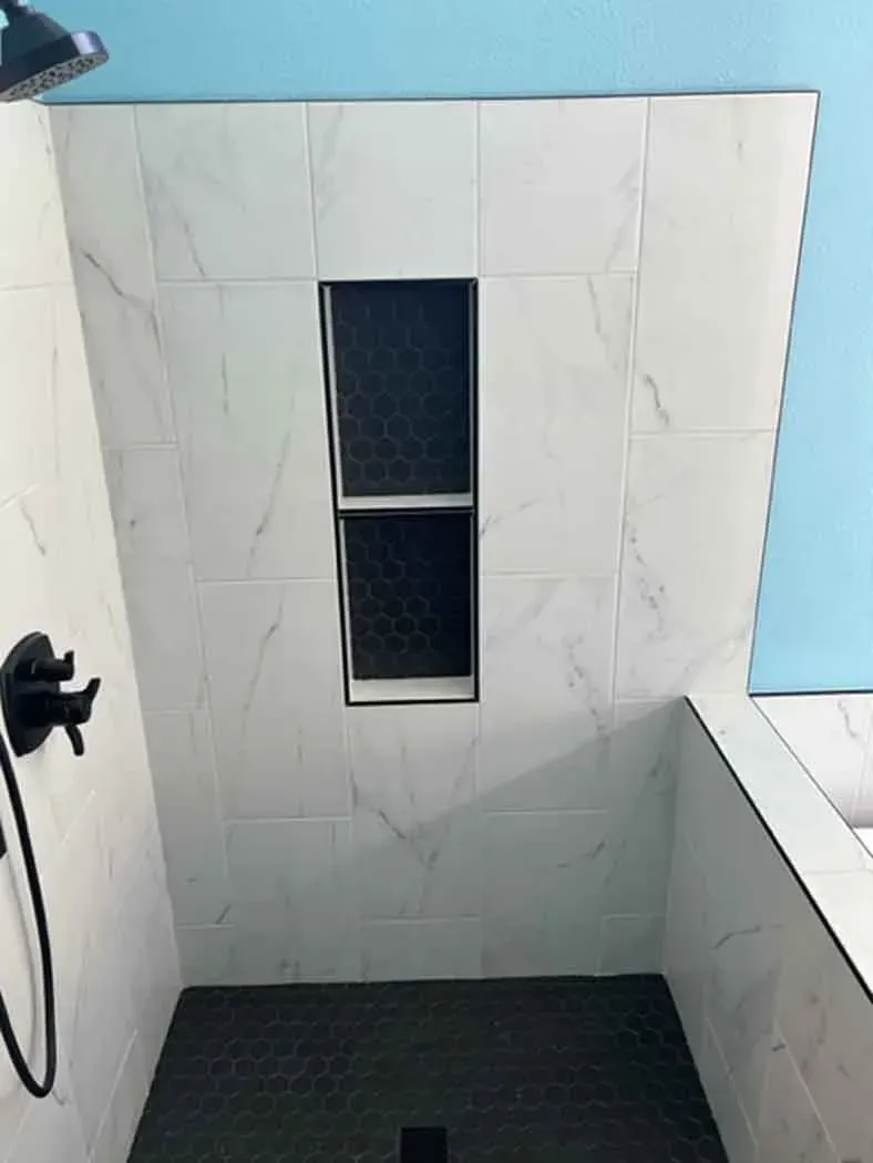 Shower with marble-patterned tile and a built-in shelf. Black hexagon floor tiles and matte black fixtures. Blue wall.