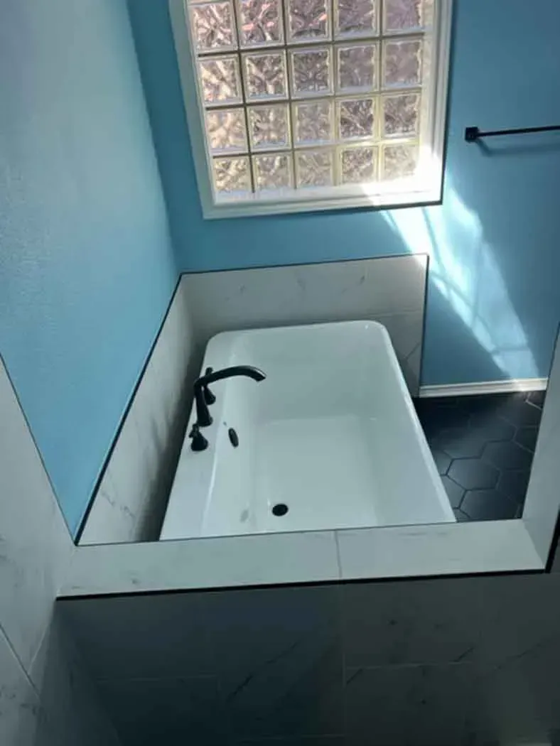 White bathtub in a blue-walled bathroom with dark faucet. Window above, black tile floor.