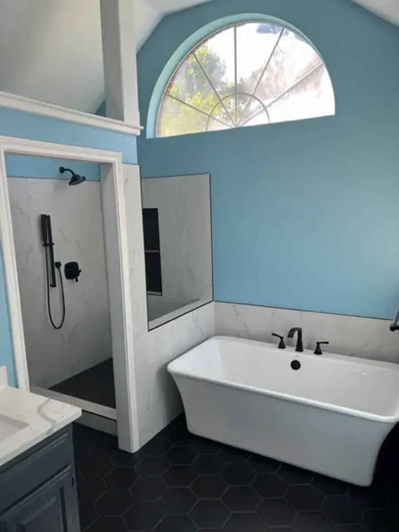 Bathroom with a white tub, shower, and light blue walls. Black fixtures and dark hexagonal floor tiles.