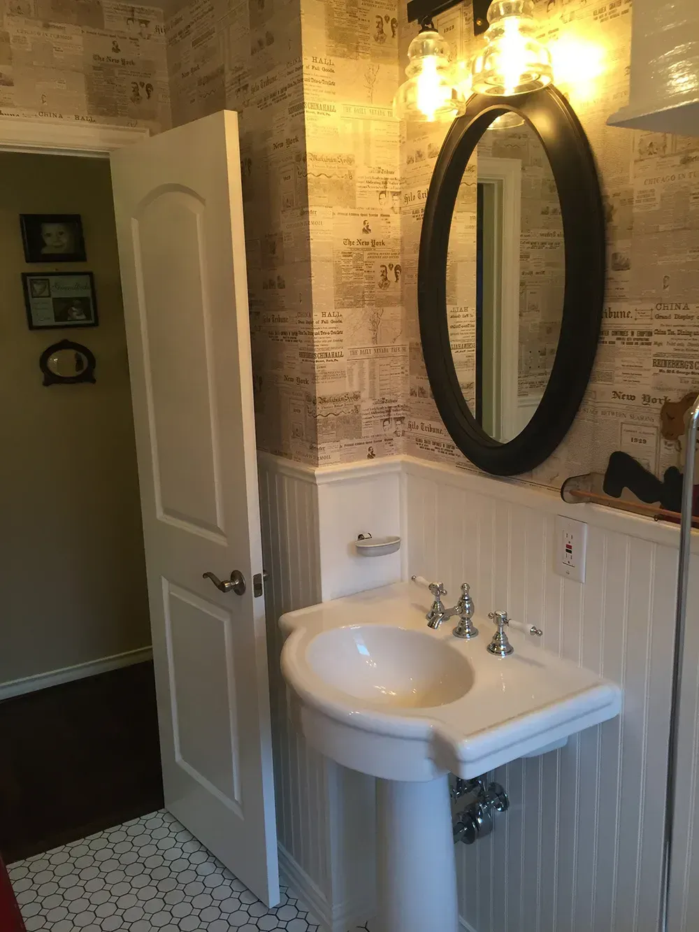 Bathroom with white pedestal sink, black oval mirror, and patterned wallpaper. Doorway on the left.