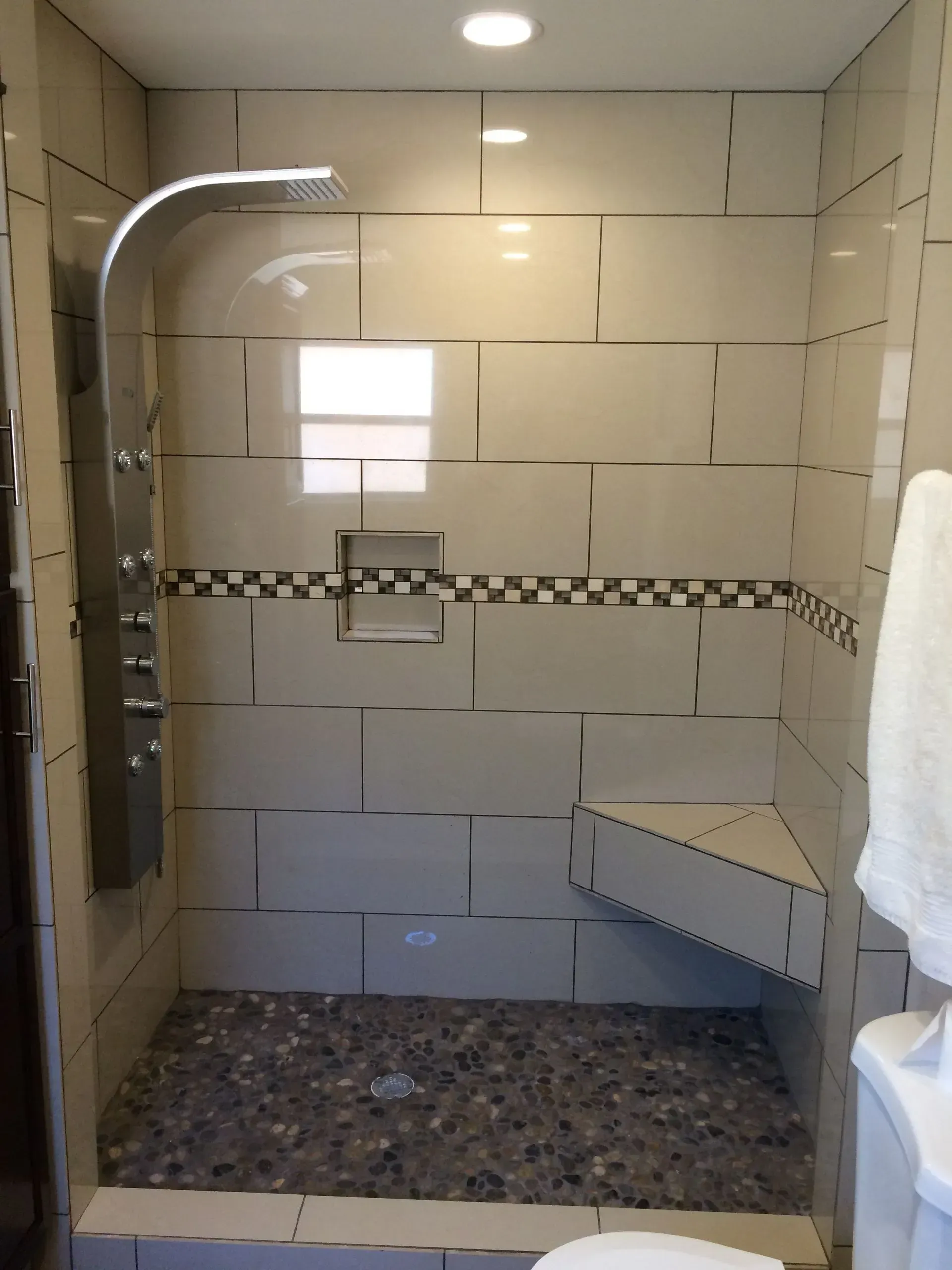Tiled shower with pebble floor, built-in bench, and rain showerhead. Beige and brown tones.