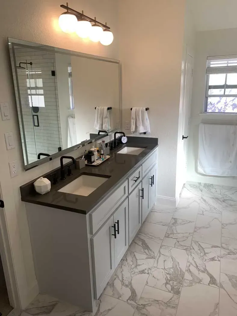 Bathroom with white cabinets, dark countertop, double sinks, large mirror, and marble-look floor.