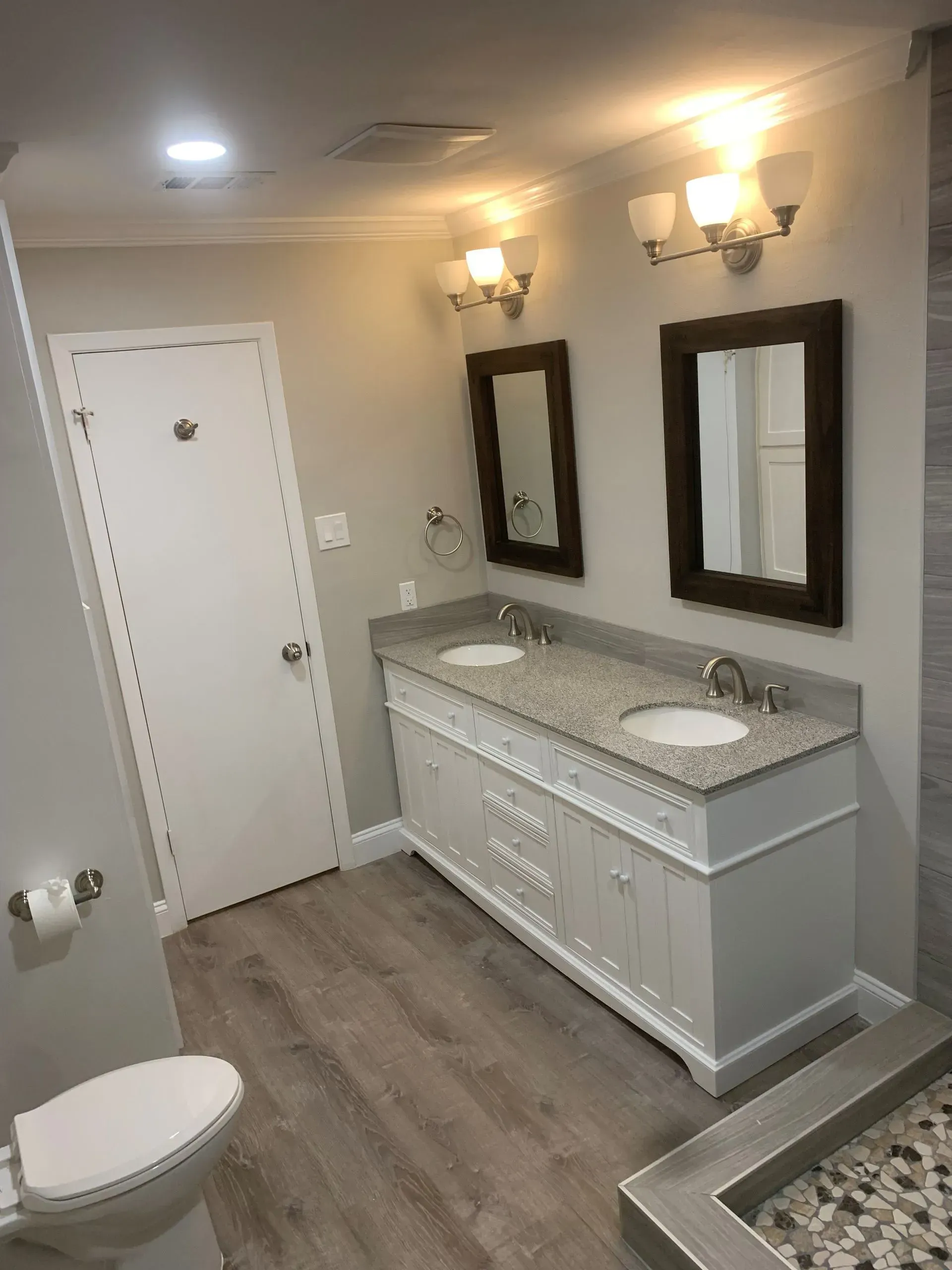 Bathroom with double vanity, two mirrors, and a toilet. Gray walls, wood-look floor, and a shower with pebble floor.