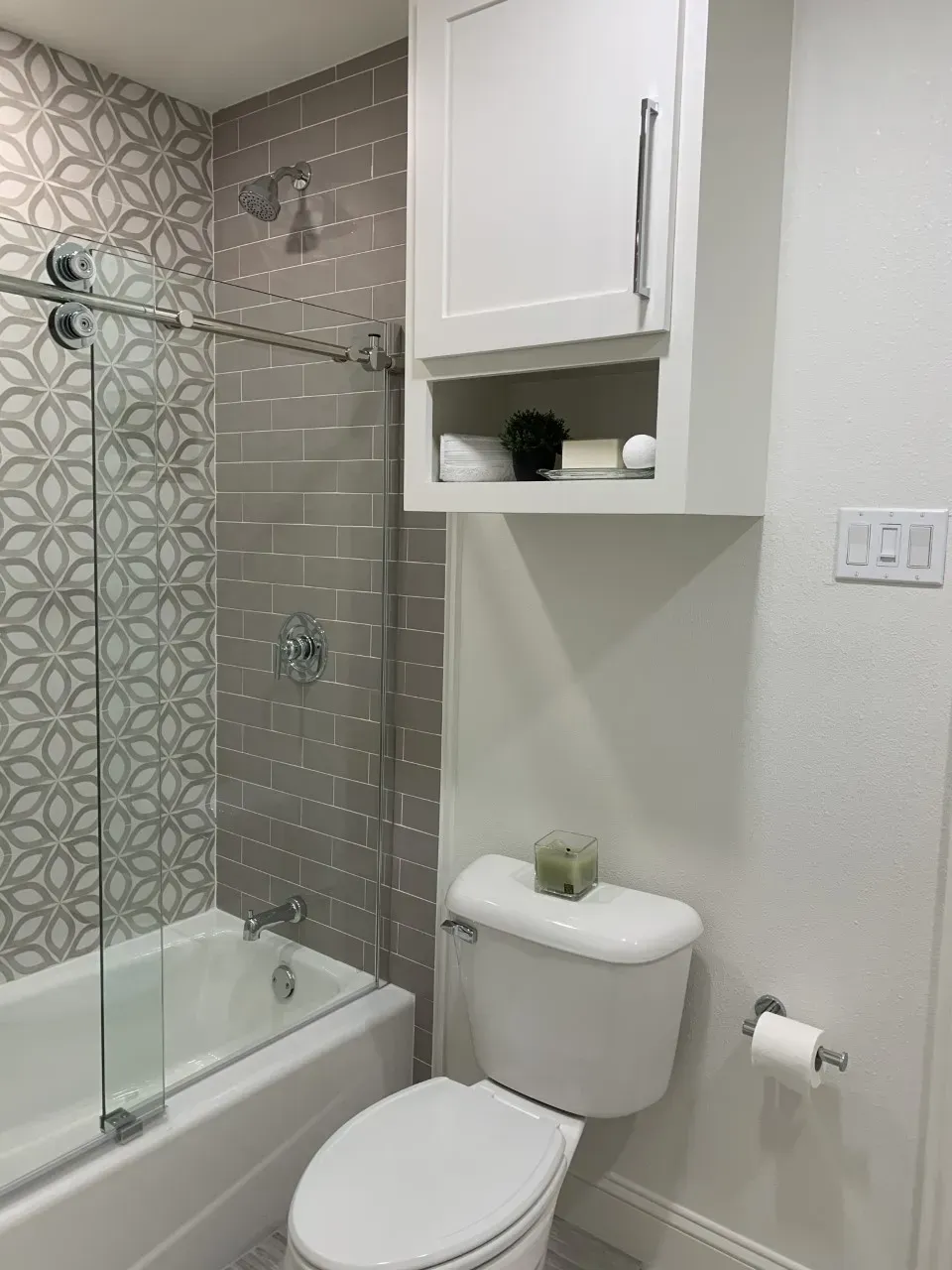 Bathroom with white walls, gray tile shower, white toilet, and cabinet.