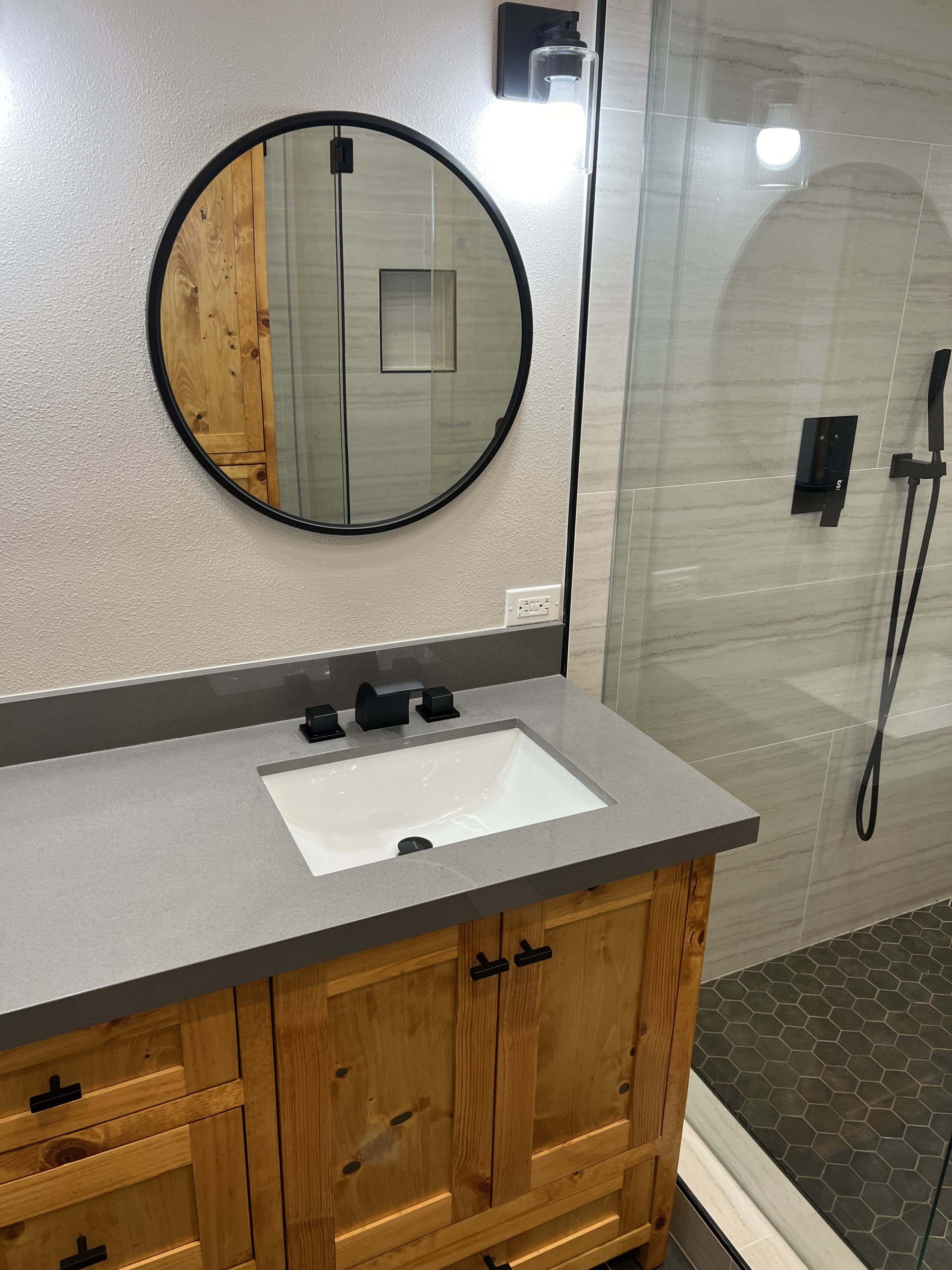 Bathroom with wooden vanity, gray countertop, black fixtures, round mirror, and glass shower.