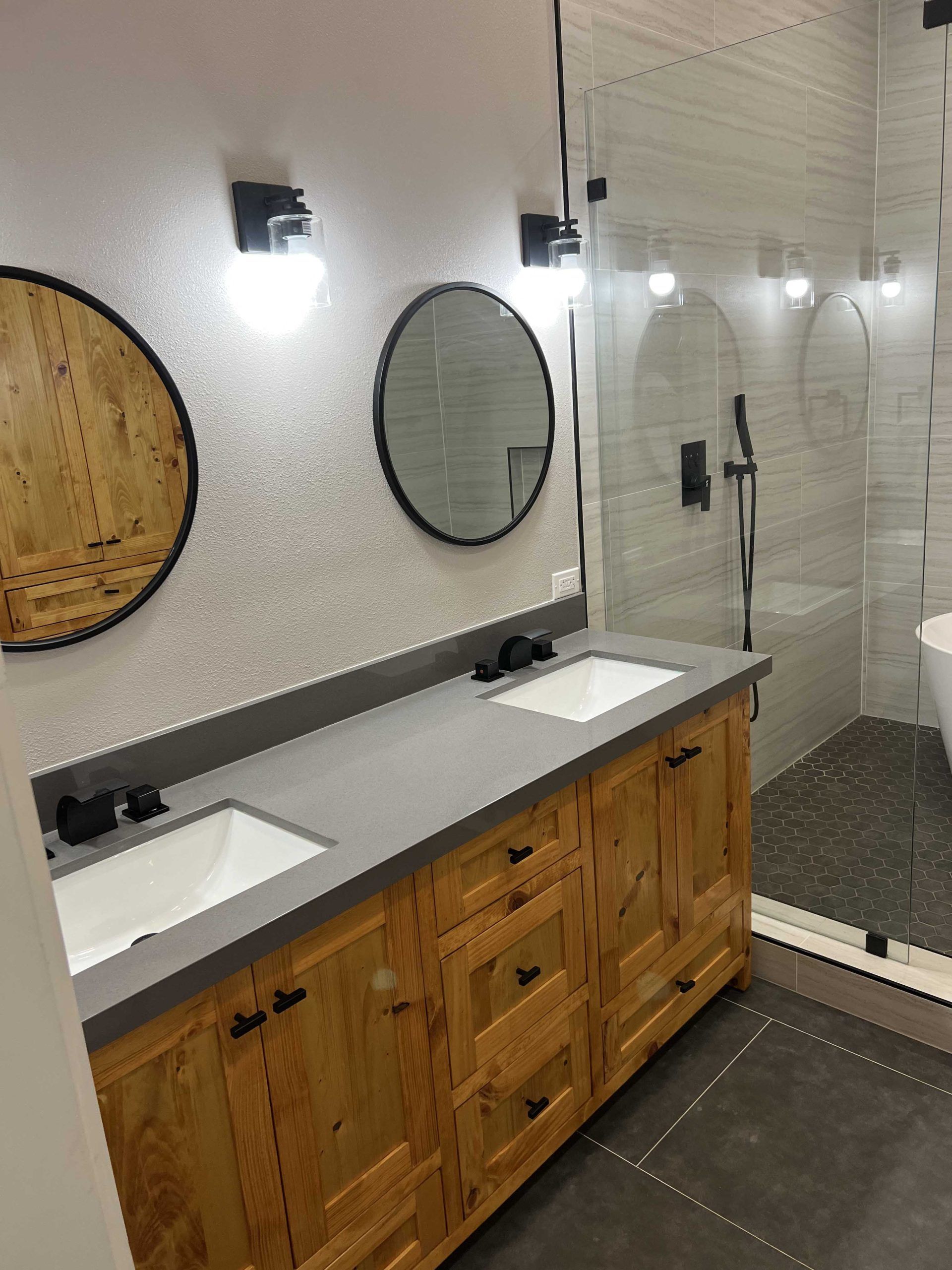 Bathroom with wooden vanity, two sinks, round mirrors, and glass shower.