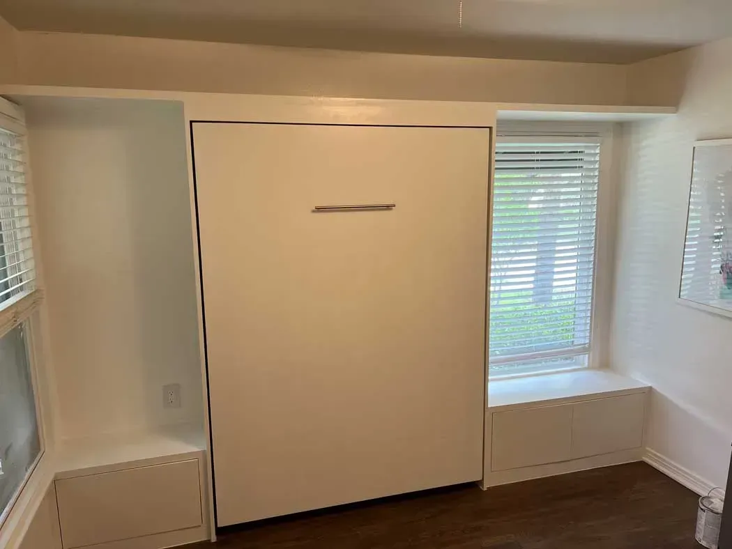 A white wall bed is closed against a window and side ledges in a room with hardwood floors.