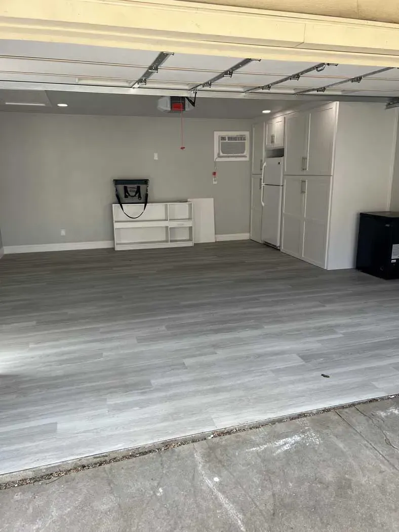 Empty garage with gray wood-look floor, white shelves, and cabinets.