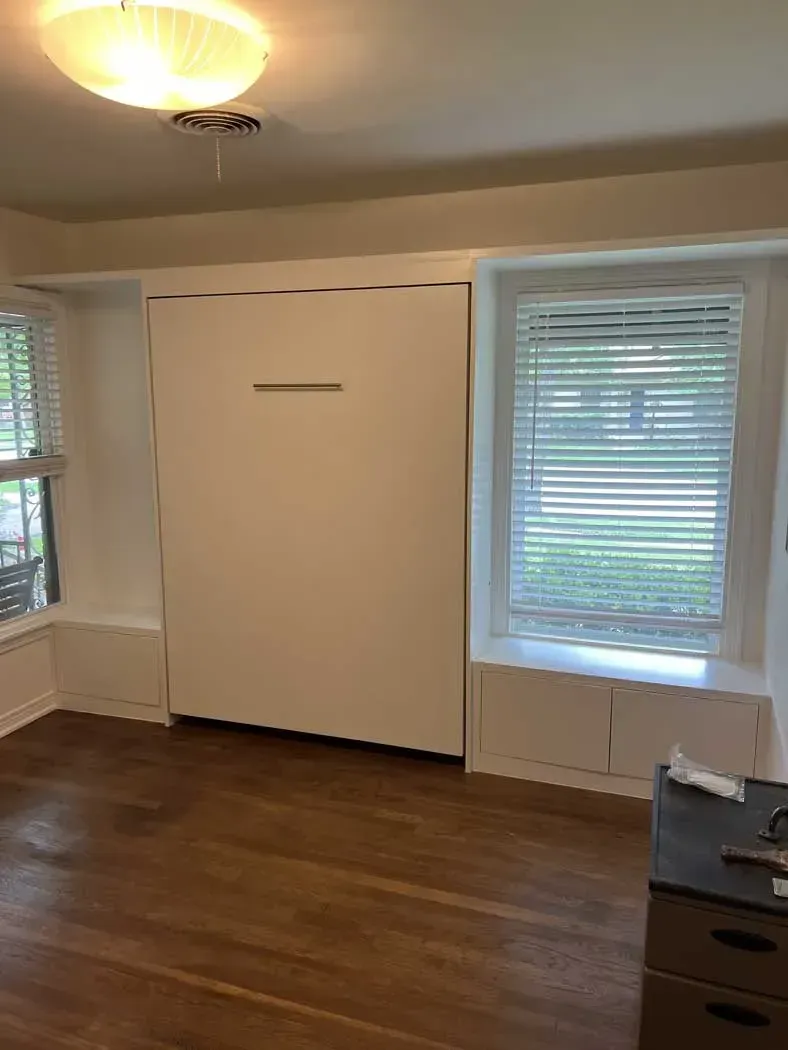 A white wall bed is closed between two windows. Hardwood floor and a light fixture visible.