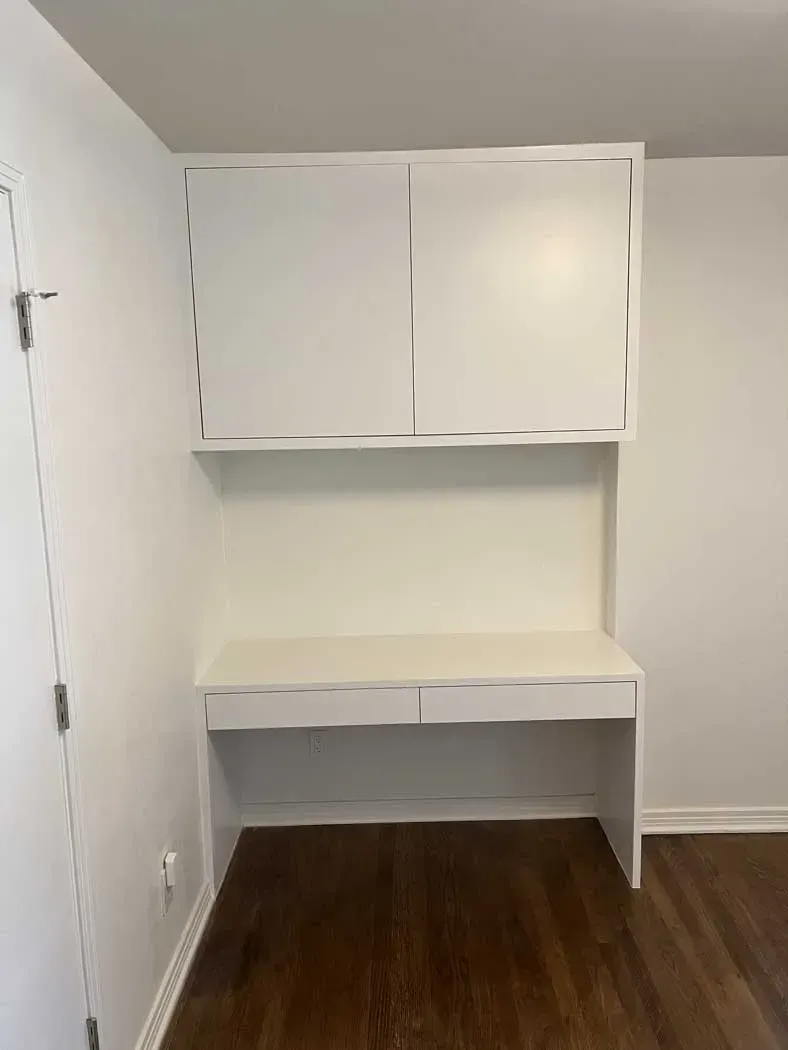 Built-in white desk and upper cabinets in a corner of a room, against white walls and wood flooring.