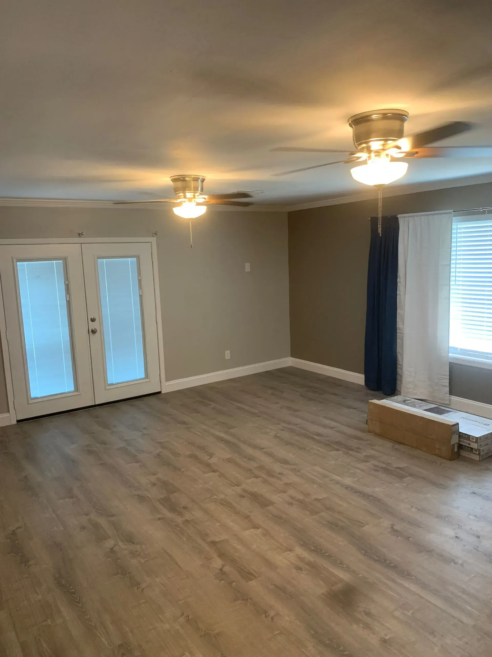 Empty living room with wood-look flooring, beige walls, two ceiling fans, and a double door leading outside.