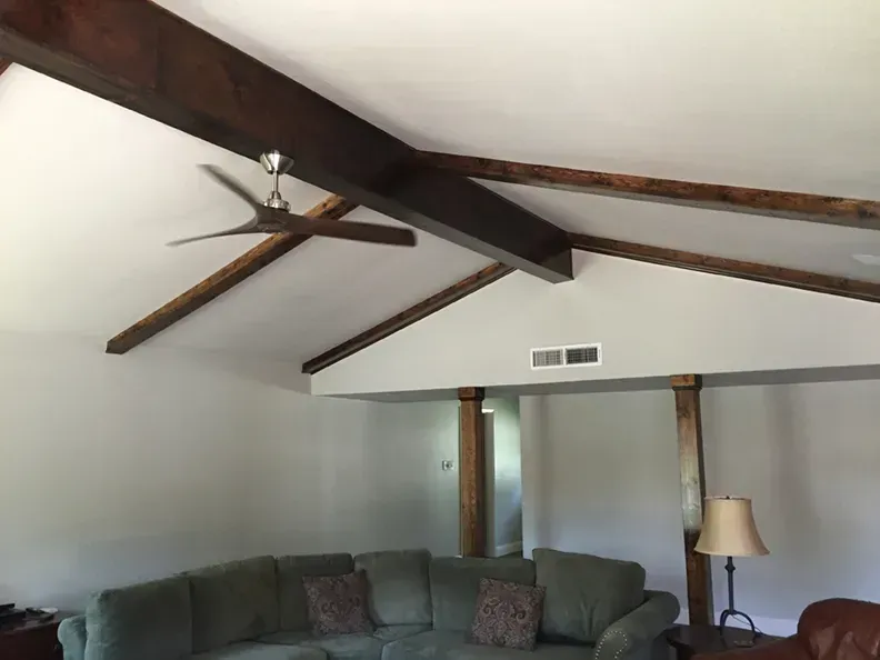 Living room with angled ceiling, exposed dark beams, and a ceiling fan. A green couch is in the foreground.