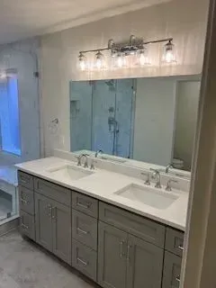 Bathroom with gray cabinets, white countertop, double sinks, large mirror, and glass shower.