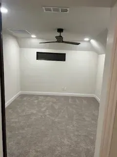Empty, white-walled room with gray carpet, ceiling fan, and small window; viewed from doorway.