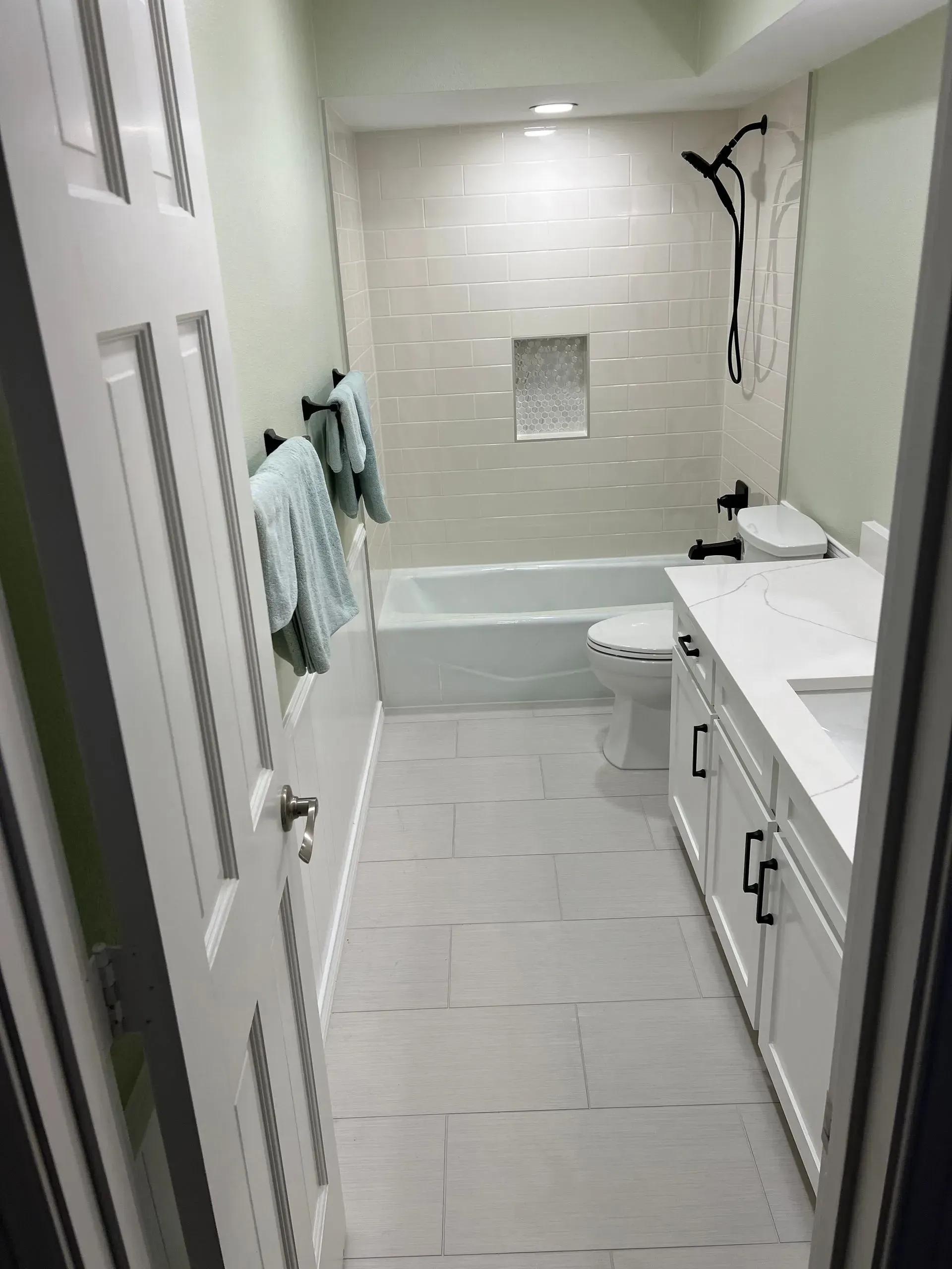 Narrow, renovated bathroom with white and light green walls, a white vanity, and tub.