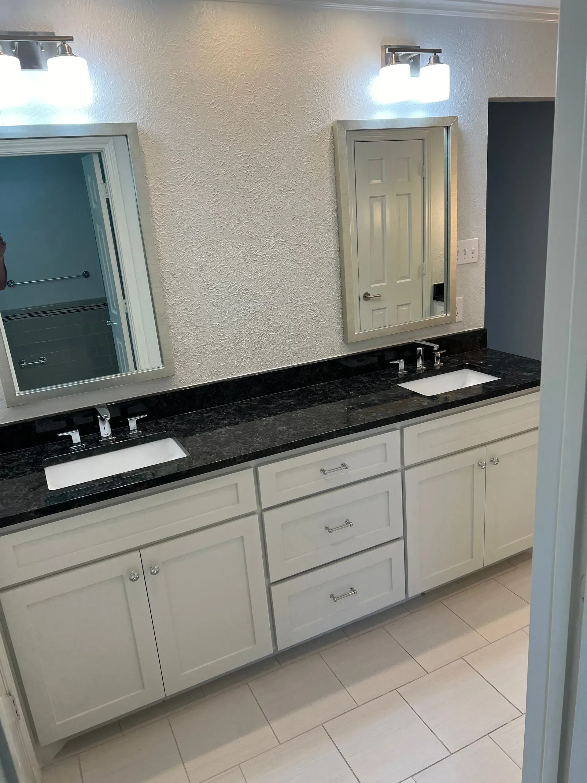 Bathroom with two sinks, mirrors, and white cabinets under a black countertop. White walls with mosaic tiles.