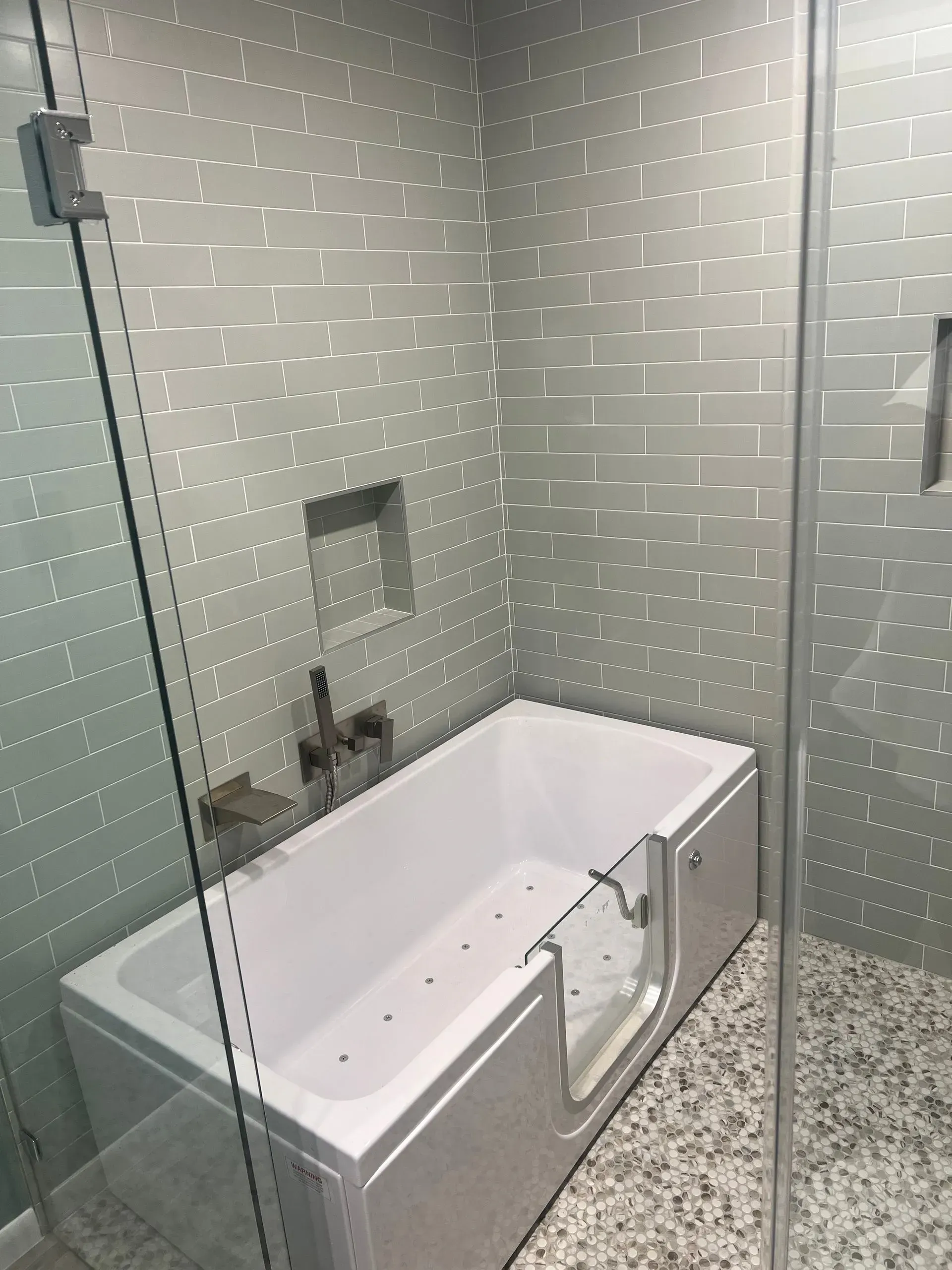 Walk-in bathtub with glass door in a bathroom with textured gray tile walls.