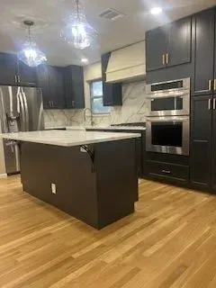 Modern kitchen with dark gray cabinets, white countertops, and wood flooring. Island in center, stainless steel appliances.