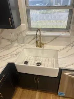 White farmhouse sink with gold faucet in kitchen, set against marble countertops and a window.