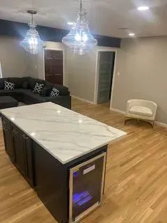 Modern kitchen with island, marble countertop, dark cabinets, wine cooler, chandelier lighting, and a grey sectional sofa.