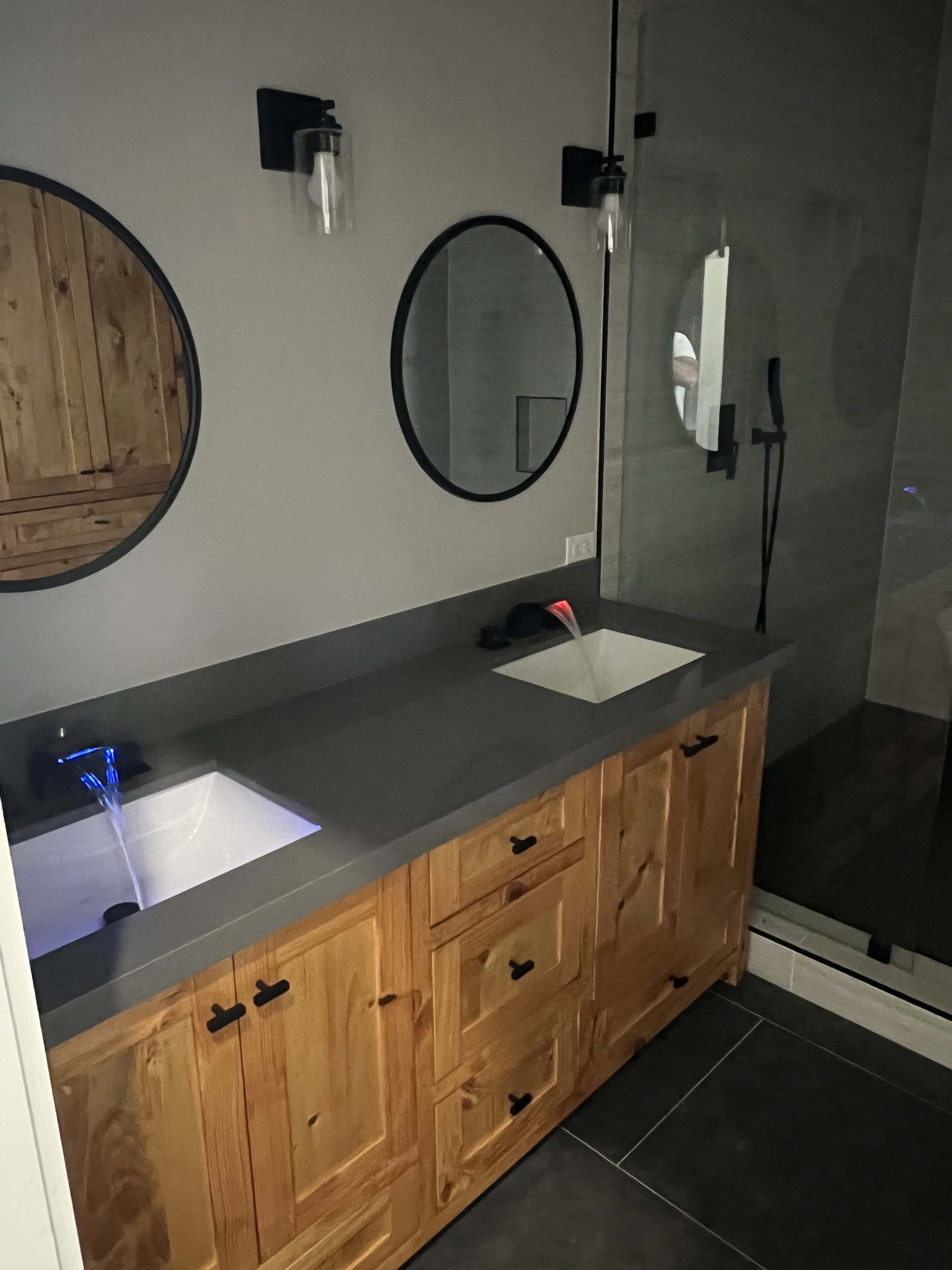 Bathroom with wooden vanity, gray countertop, round mirrors, and black sconces.