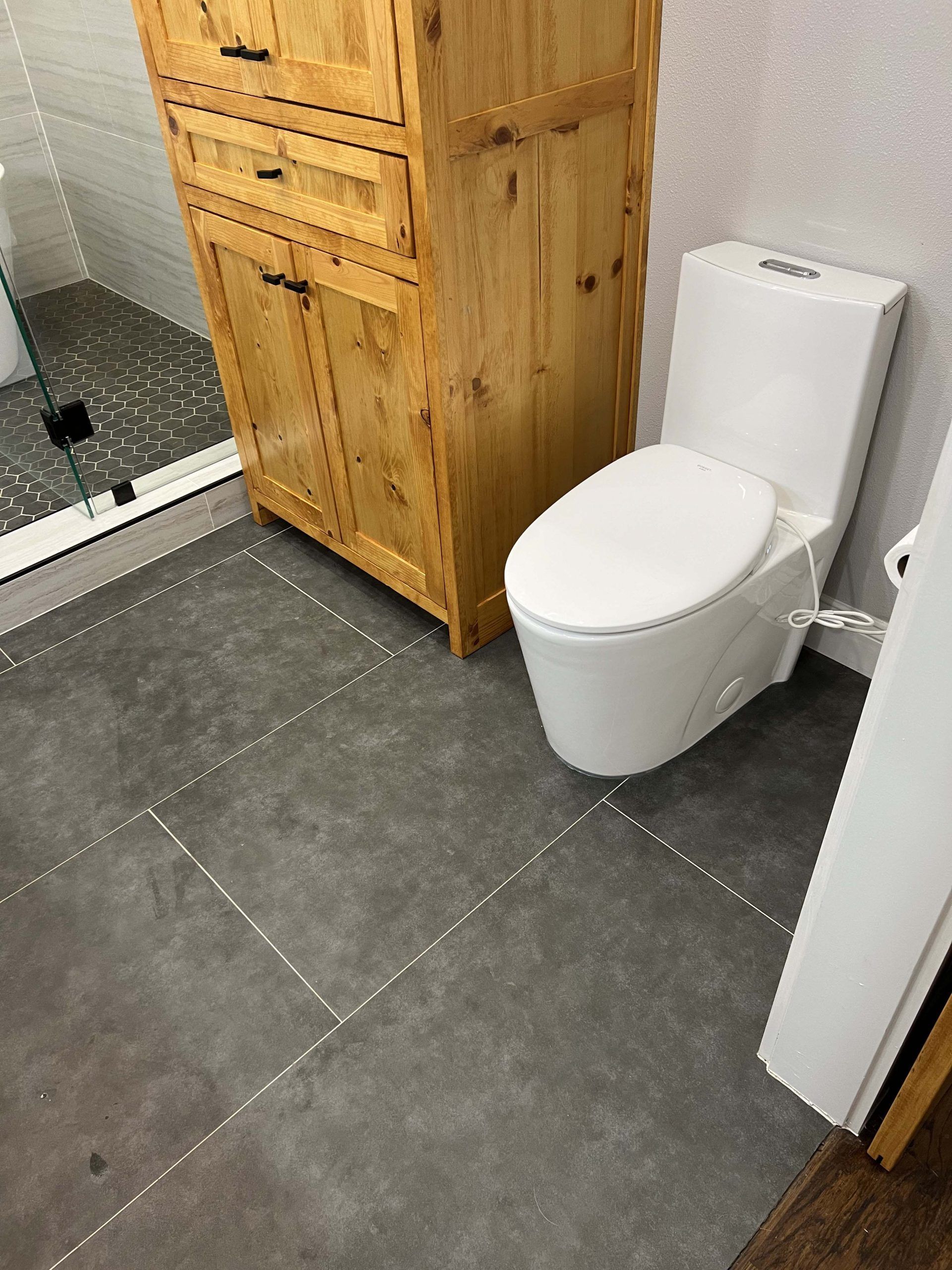 Bathroom with a white toilet, wooden cabinet, and gray tile flooring. A shower is visible on the left.