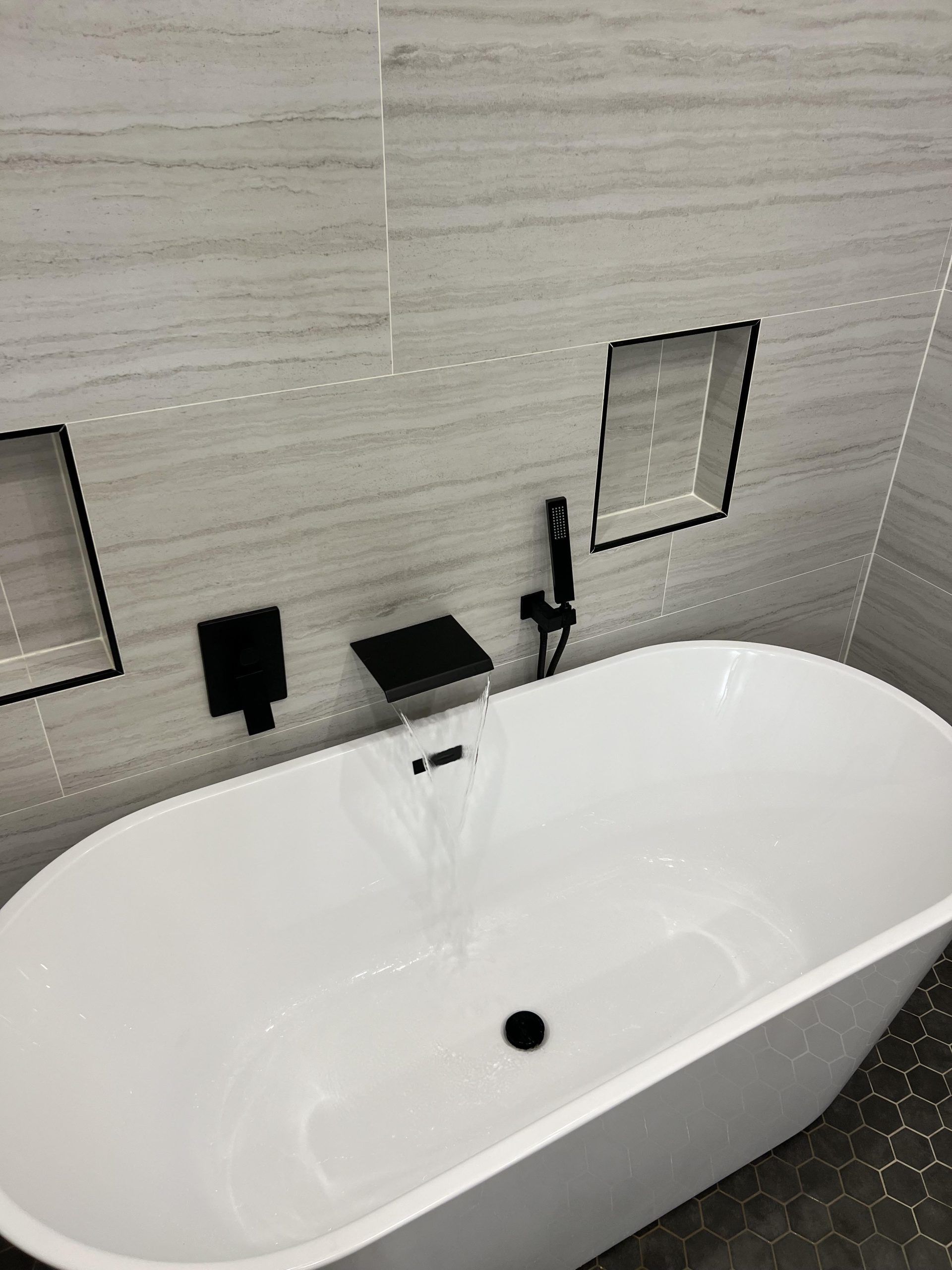 White bathtub with black fixtures against light gray tiled wall; two recessed shelves.