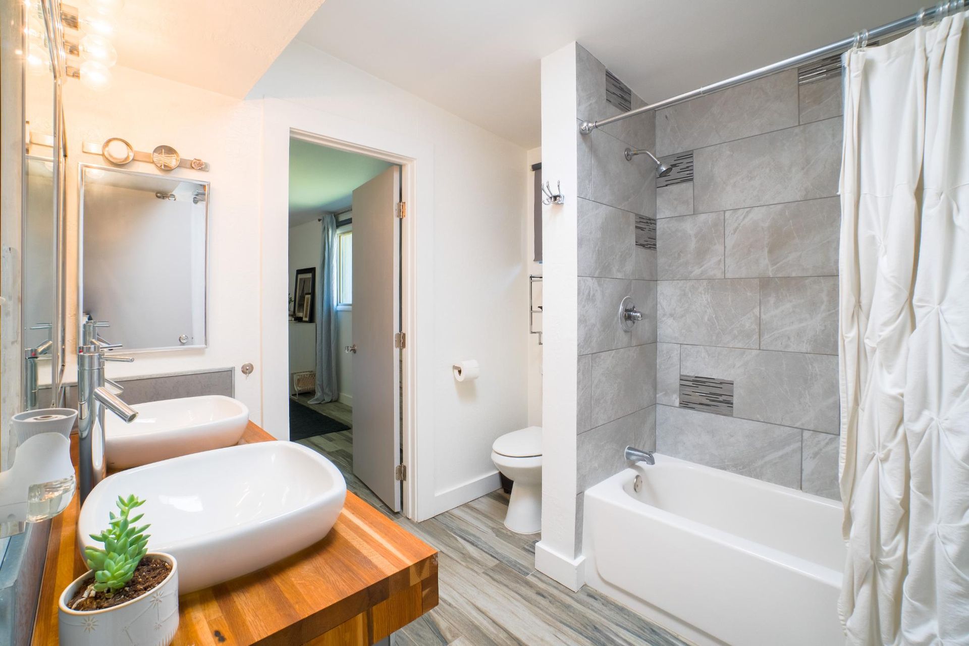Bathroom with a modern design: white sinks on a wooden counter, a white tub with gray tile, and an open doorway.
