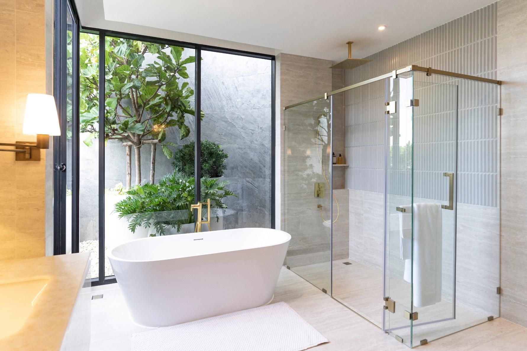 Modern bathroom with a white tub, glass shower, and view of a green garden through a large window.