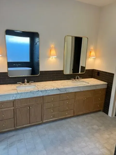 Bathroom with double vanity, gold-framed mirrors, sconces, wood cabinets, marble countertop, and dark tiled backsplash.