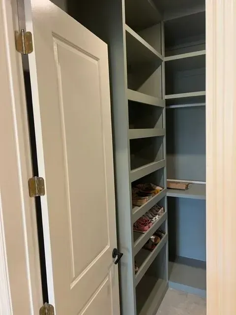 A walk-in closet with shelves and a shoe rack, painted in a neutral green color; open door with brass hinges.