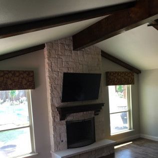Living room with stone fireplace, TV, dark wood beams, and patterned window valances.
