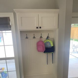 White built-in entryway storage with cabinets, hooks, and hanging backpacks. Next to a window and doorway.