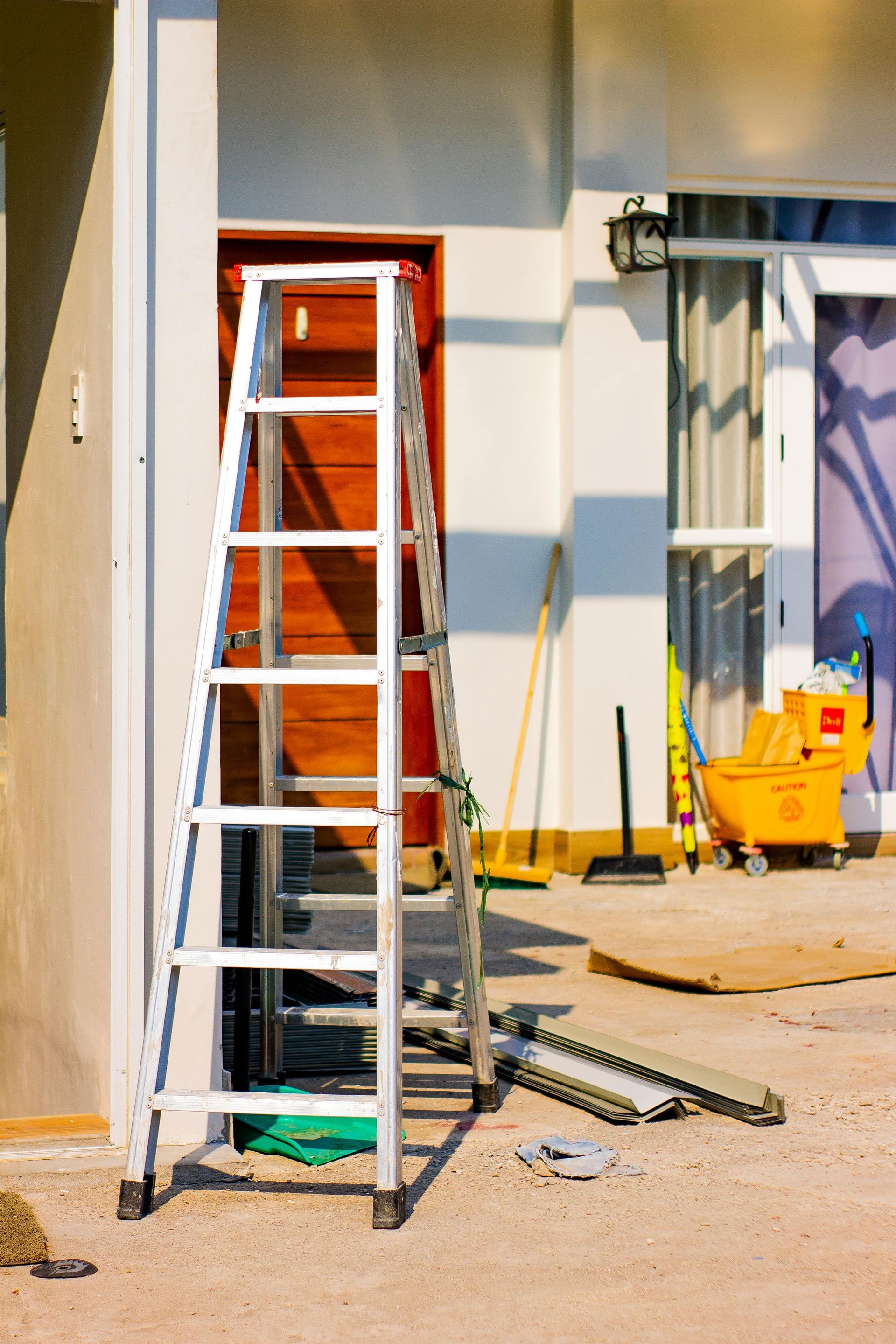 Aluminum ladder propped against a building with a red door. Construction materials and tools scattered nearby.