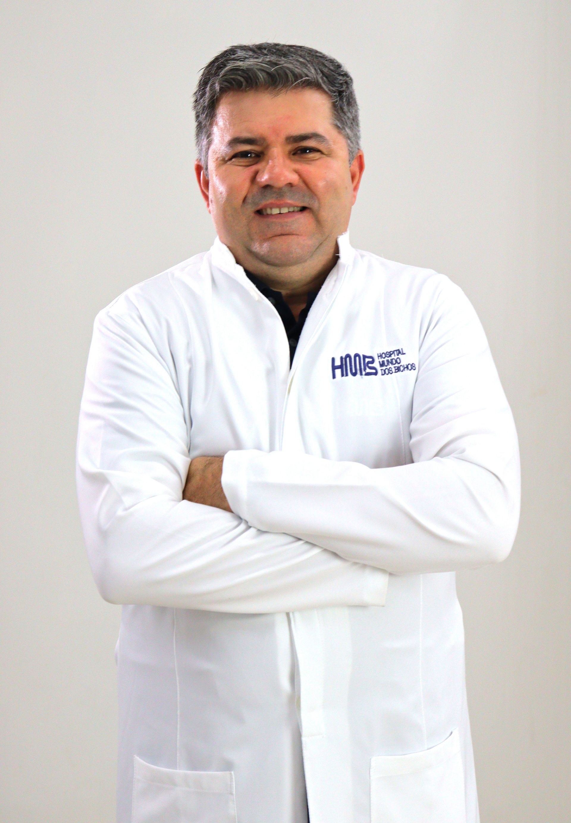 Homem de jaleco branco, com os braços cruzados, sorrindo. Placa do hospital ao fundo.