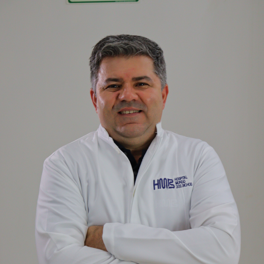 Homem de jaleco branco, com os braços cruzados, sorrindo. Placa do hospital ao fundo.