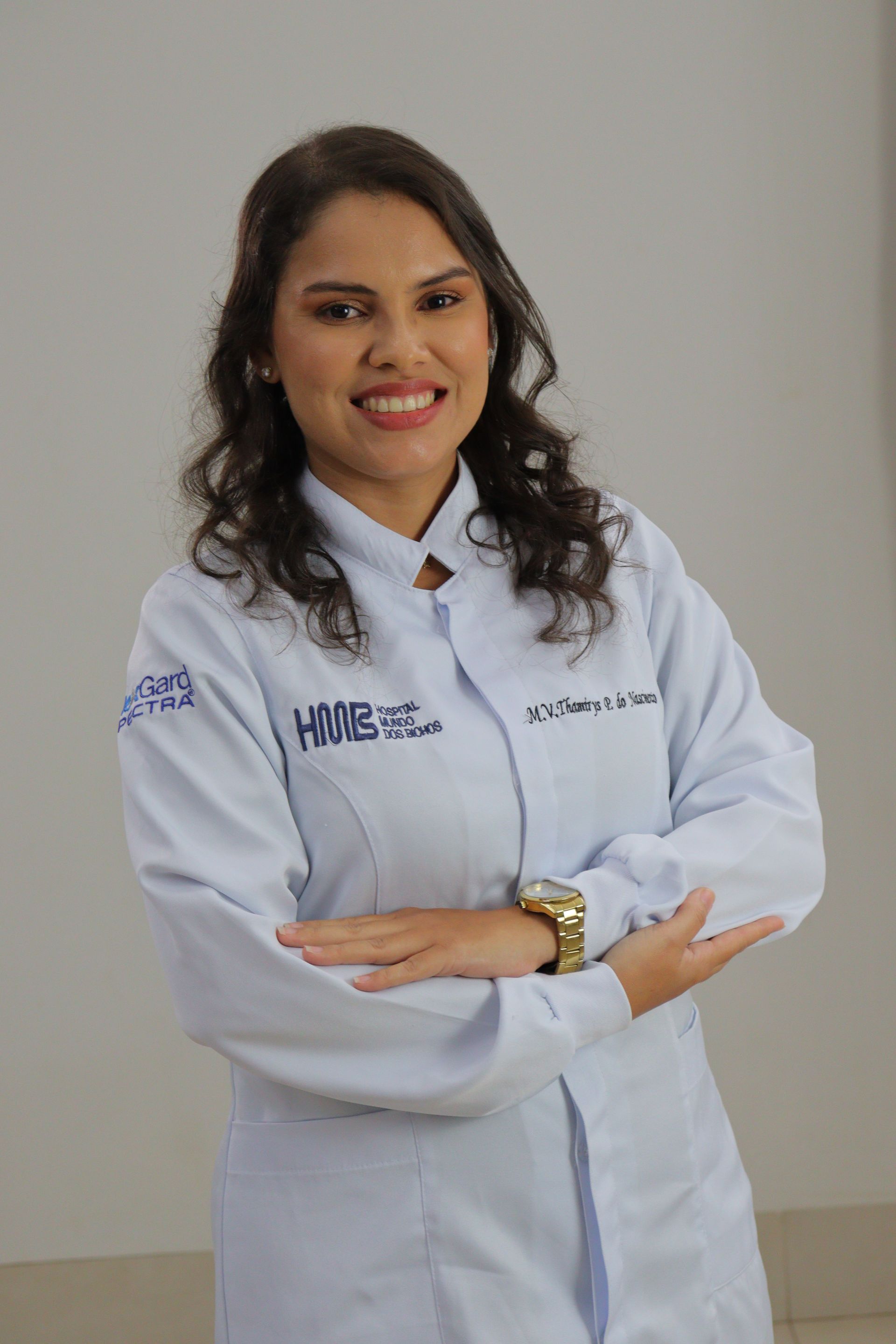 Mulher vestindo uniforme cirúrgico verde-azulado e estetoscópio, braços cruzados, sorrindo.