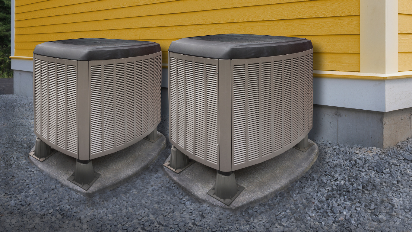 Two beige and black air conditioning units sit outside a yellow house on concrete pads.