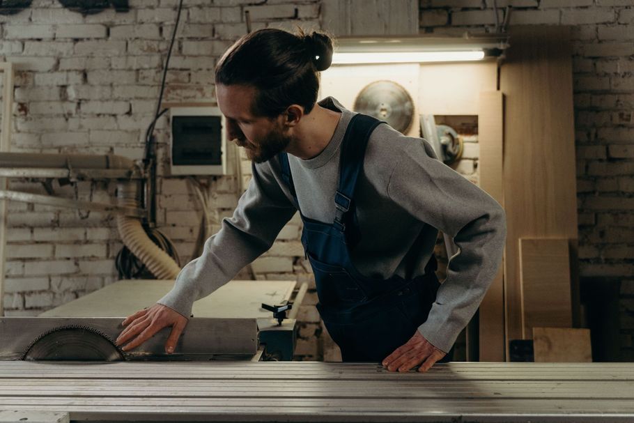 Worker using a table saw in a workshop, leaning over wood with a focused expression.