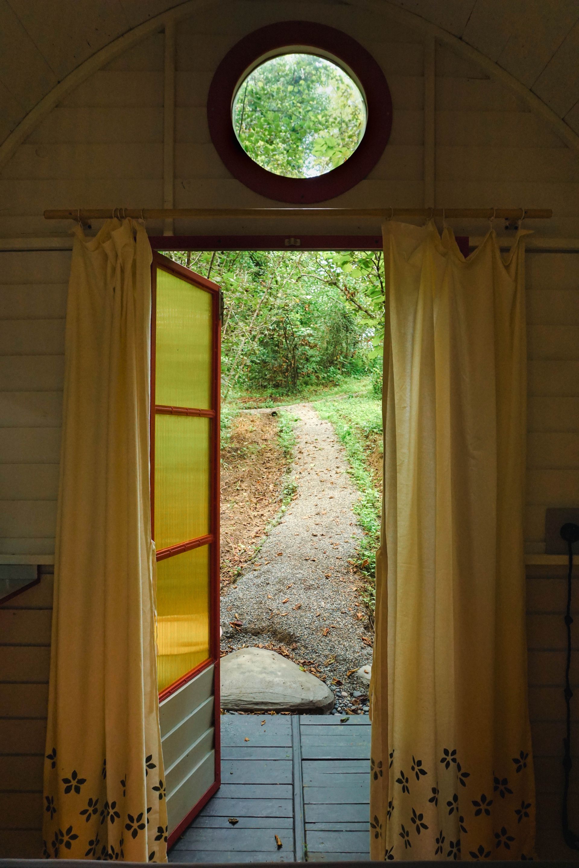 A door with a round window in it is open to a path in the woods.