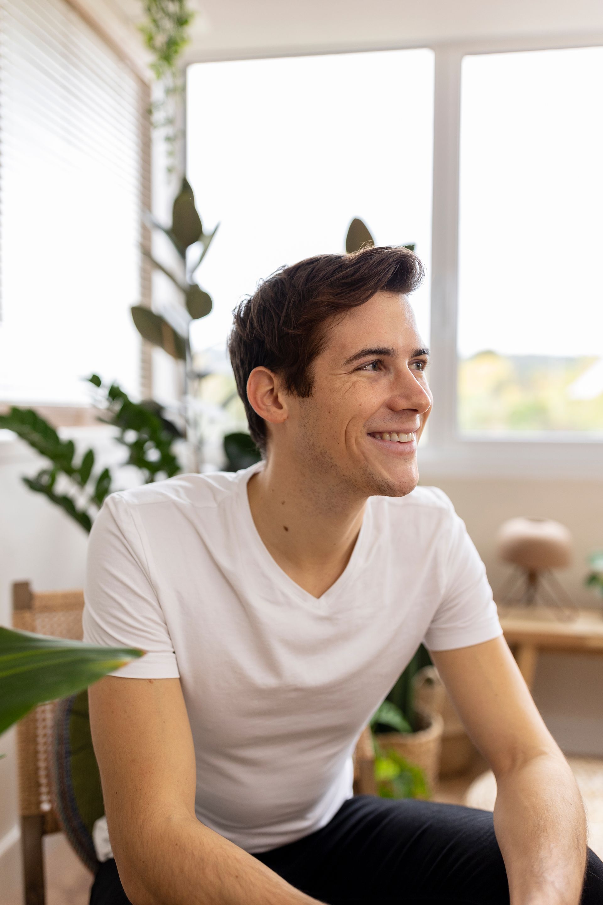 A man in a white t-shirt is sitting in a chair in front of a window.