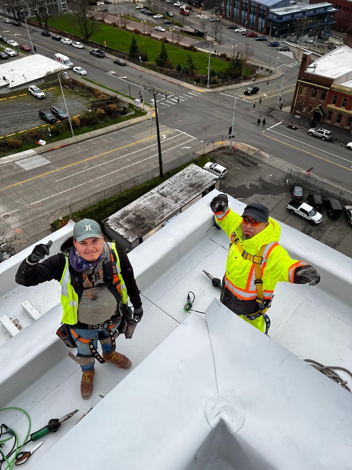 Two workers on a rooftop, one in a vest. City view below.
