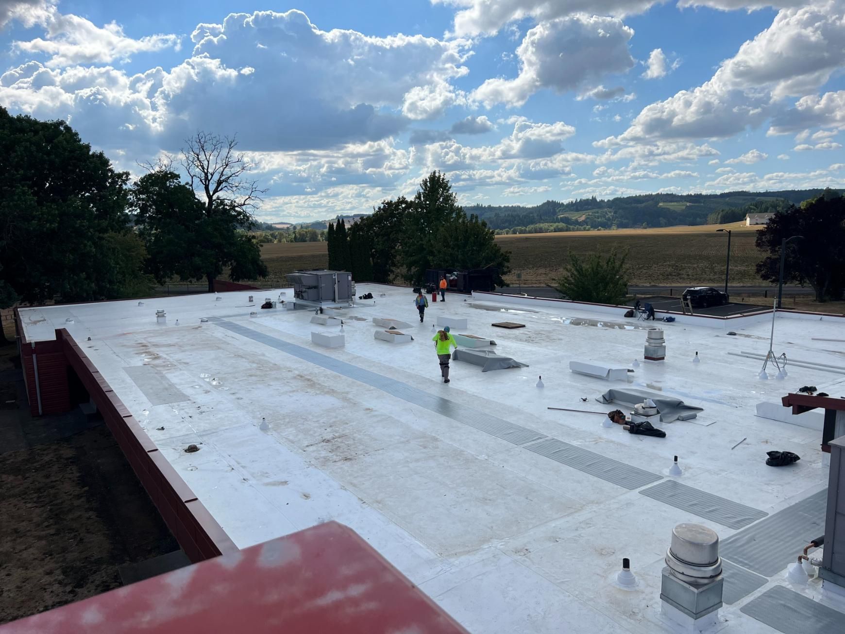 Workers on a flat roof with white coating, surrounded by trees and fields under a cloudy sky.
