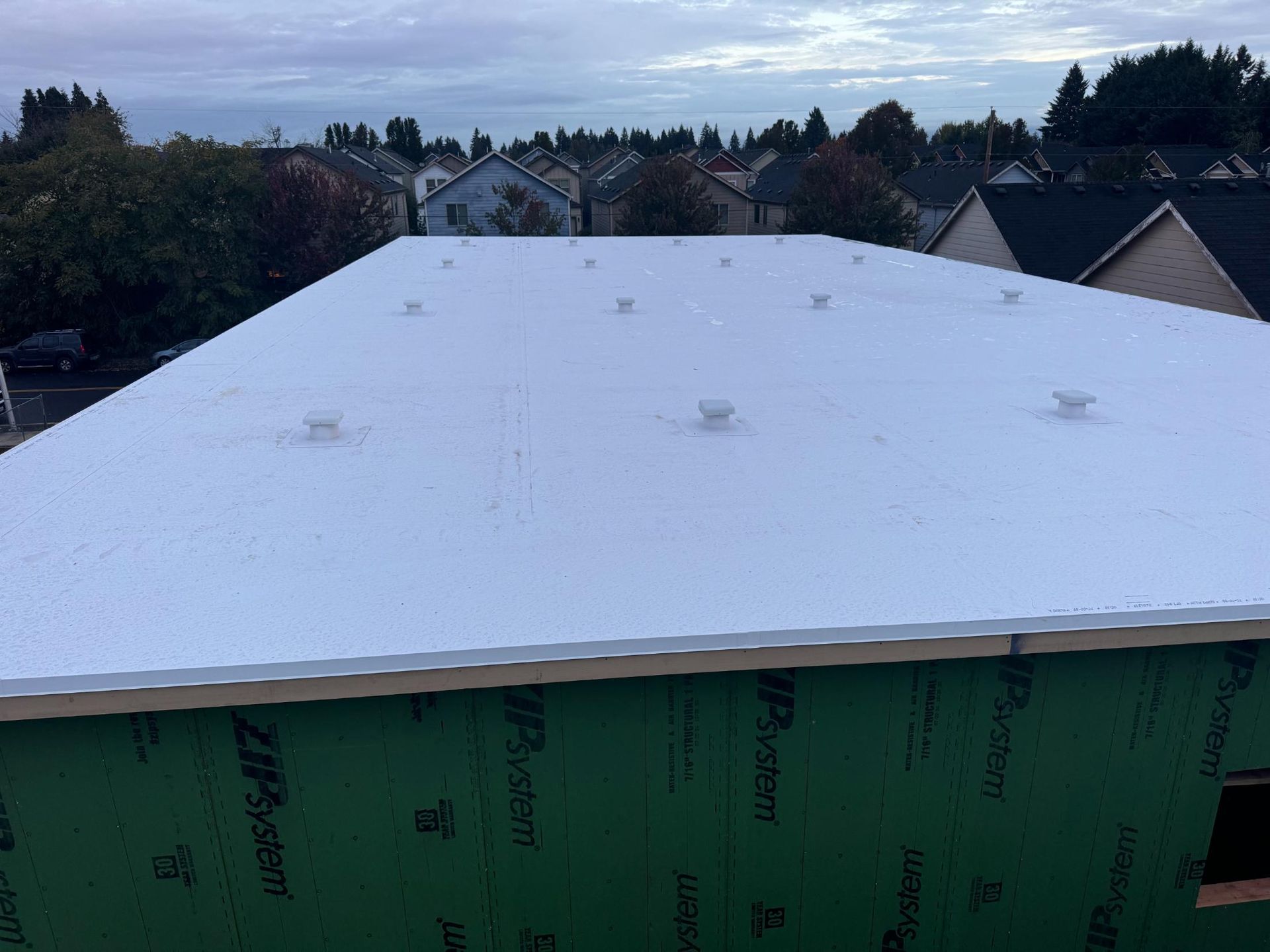 White flat roof on a partially built building, with green sheathing and vents visible. Houses and trees in background.